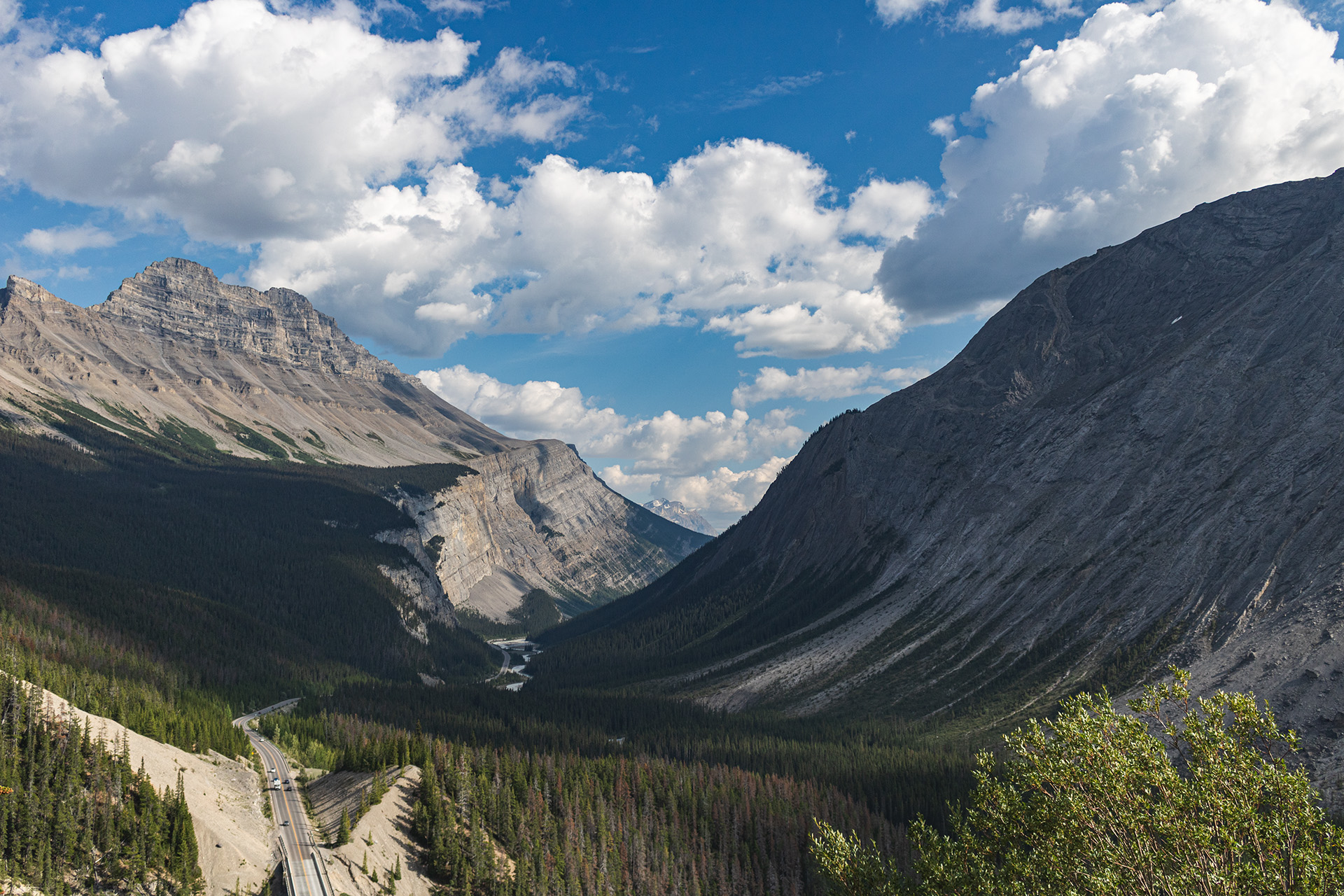 Valley with a giant sweeping rocky mountain on each side and a road passing below, seen from halfway up the mountains' height.